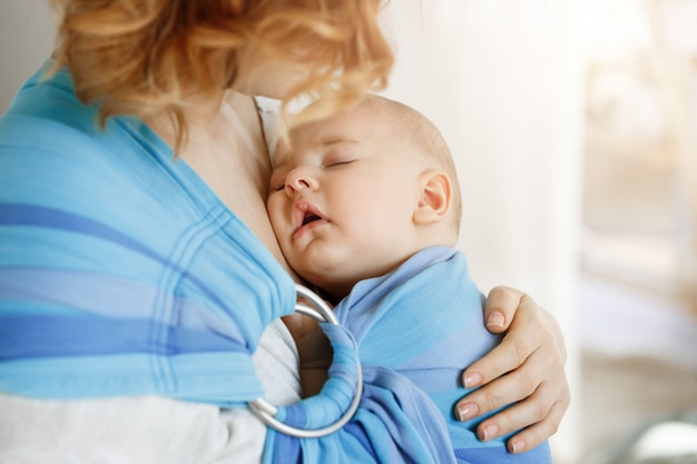Close up portrait of innocent newborn boy having sweet dreams on mother chest in baby sling. Mom looking at her child with love and tenderness.