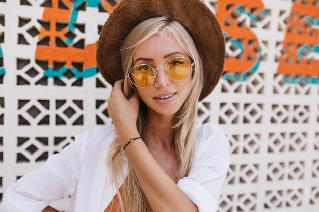 Ritratto del primo piano della donna bionda ispirata con capelli lunghi che posano nel giorno di estate. foto del bellissimo modello femminile interessato in cappello marrone.