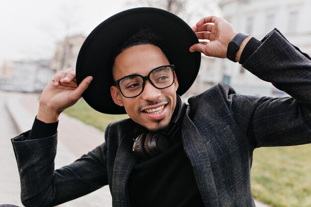 Close-up portrait of inspired man with brown skin wears casual glasses. Outdoor shot of young african male model in black attire.