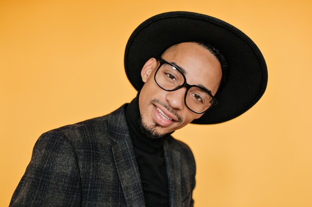 Close-up portrait of interested african guy in trendy glasses. Indoor photo of enchanting black man smiling on orange wall.