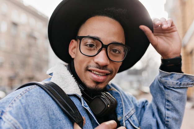 관심이 아프리카 남자의 클로즈업 초상화는 우아한 모자를 착용합니다. 데님 재킷에 잘 생긴 흑인 남자의 야외 사진.