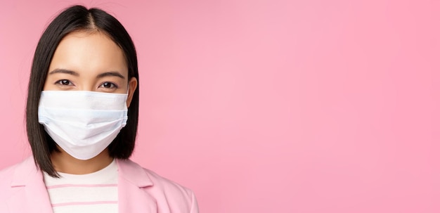 Close up portrait of japanese businesswoman in medical face mask suit looking at camera standing ove