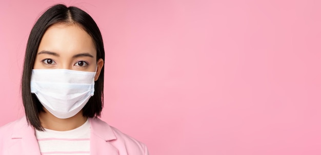 Free photo close up portrait of japanese businesswoman in medical face mask suit looking at camera standing over pink background