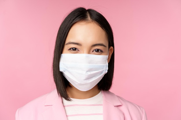 분홍색 배경 위에 서 있는 카메라를 바라보는 의료용 얼굴 마스크 정장을 입은 일본 여성 사업가의 초상화를 클로즈업