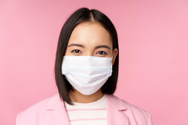 Close up portrait of japanese businesswoman in medical face mask suit looking at camera standing over pink background