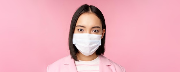 ピンクの背景の上に立っているカメラを見て医療フェイスマスクスーツで日本の実業家の肖像画をクローズアップ