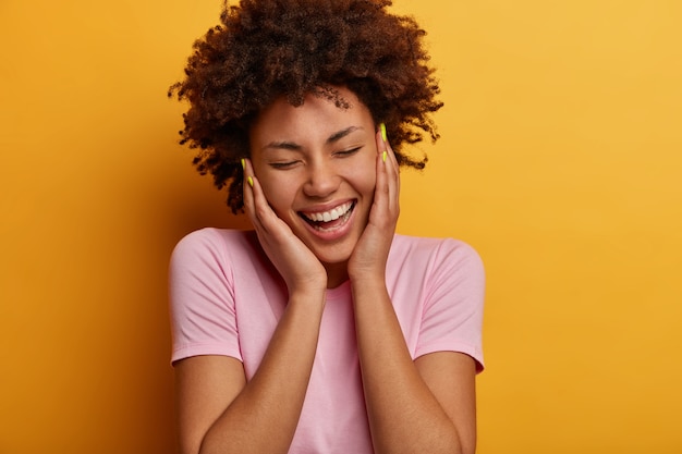 Close up portrait of joyful curly haired woman touches cheeks, has sincere positive smile, closes eyes and being amused, laughs from something hilarious, dressed casually, isoalated over yellow wall