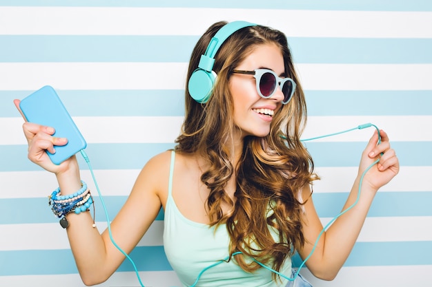 Free photo close-up portrait of joyful girl enjoying music in big headphones, holding mobile phone in hand. attractive young woman wearing black sunglasses and trendy accessories chilling on striped wall.