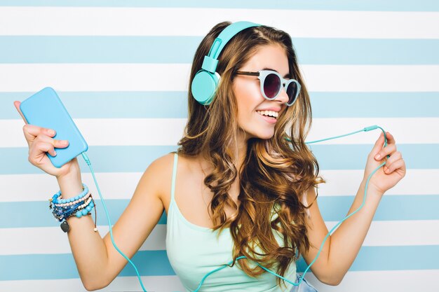 Close-up portrait of joyful girl enjoying music in big headphones, holding mobile phone in hand. attractive young woman wearing black sunglasses and trendy accessories chilling on striped wall.