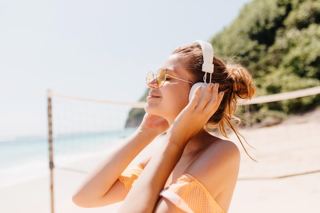 Close-up portrait of joyful tanned woman relaxing with favorite music at beach. Outdoor shot of smiling female model in headphones spending time at resort.