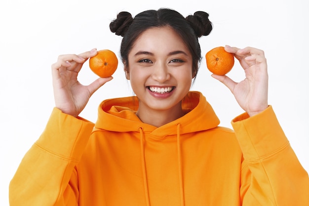 Ritratto ravvicinato di una giovane ragazza asiatica sorridente kawaii con due mandarini, risatina sciocca e guardando la macchina fotografica, mangiando frutta, giocando con i mandarini, scherzando sul muro bianco e infantile