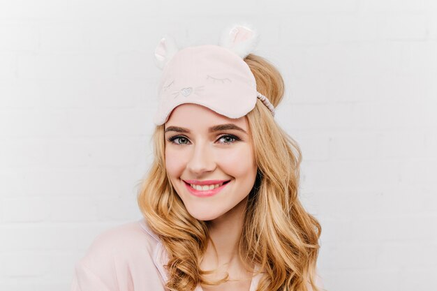 Close-up portrait of laughing gorgeous woman in cute eyemask. Pleased european female model in pajama enjoying good morning on white wall.