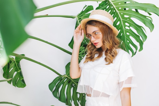 Close-up portrait of lightly tanned girl in elegant hat looking with serious face expression to camera. Pretty young woman in stylish attire with blonde curls posing on white background with plant.