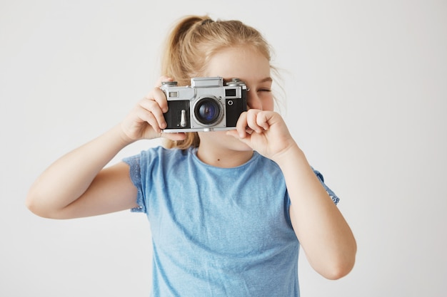 Close up portrait of little adorable girl with blonde hair in blue t-shirt going to take a picture of friends in school with film camera.