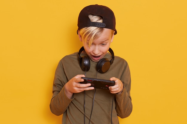 Close up portrait of little blond boy posing with mobile phone in hand isolated over yellow