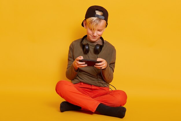 Close up portrait of little blond guy wearing casual clothing, posing with headphones around neck, playing online video games via mobile phone, looks concentrated, isolated on yellow.