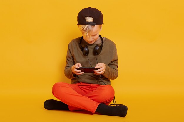 Close up portrait of little boy sitting on floor in with crossed legs isolated on yellow wall, watching video online on smartphone or playing game. Childhood and technology concept.