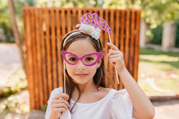 ピンクのメガネと黒髪の白いリボンの小さなファッショナブルな女性のクローズアップの肖像画。おもちゃの輝きの王冠が正面の木製のフェンスでポーズをとっている女の子の屋外写真。