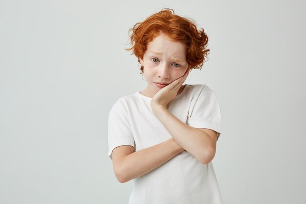 Close up portrait of little ginger boy holding head with hand looking aside with sad expression