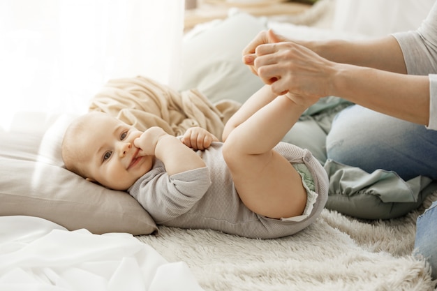 Close up portrait of little newborn son lying on bed, while playing with mother. Kid smiling and put his fingers in mouth lookin happy and carefree.