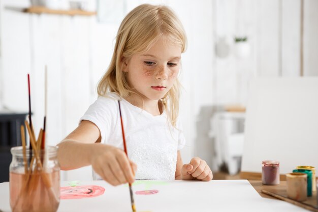 Close up portrait of little white-skinned girl with blonde hair and freckles focused on painting picture