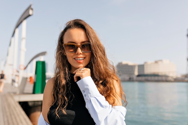 Close up portrait of longhaired european model in sunglasses and blue shirt posing at camera on background of lake Young attractive caucasian woman laughing full mouth swinging hair outdoors