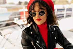 Free photo close up portrait of lovable cute lady with red lips and wearing sunglasses is smiling while waiting for friends in open air cafeteria