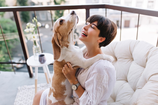 Close-up portrait of lovely black-haired lady looking with smile at funny puppy while sitting on balcony. Stunning girl in bathrobe wears bracelet and wristwatch playing with beagle dog