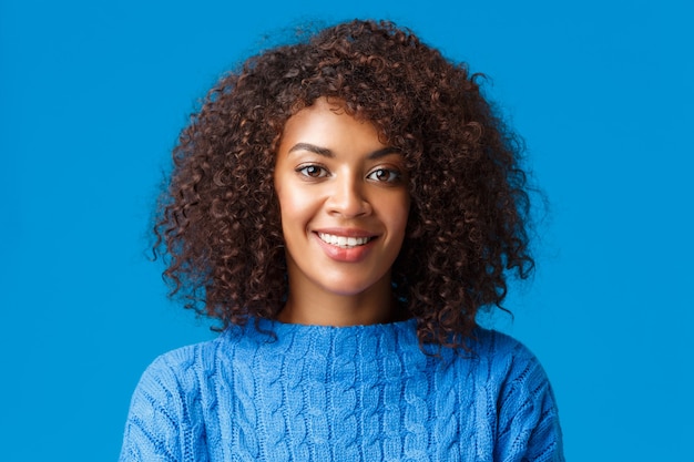 클로 우즈 업 초상화 사랑스러운 젊은 아프리카 계 미국인 여자 곱슬, 아프로 머리, 행복 한 즐거운 표정으로 웃 고, 겨울 휴가를 즐기고, 스웨터, 파란색 벽을 입고.
