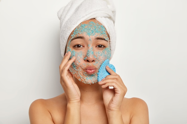 Close up portrait of lovely young female looks with folded lips, makes purity peeling procedure, has towel on her head, applies sea salt granules on face, holds sponge for wiping skin