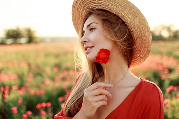 Close up portrait of lovely young romantic woman with poppy flower in hand posing on field background. Wearing straw hat. Soft colors.