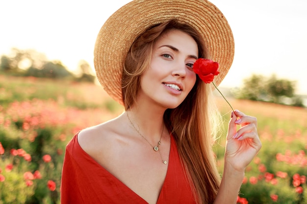 Close up portrait of lovely young romantic woman with poppy flower in hand posing on field background. Wearing straw hat. Soft colors.