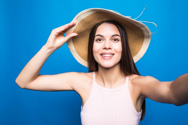 Close up portrait of a lovely young woman in summer dress and straw hat taking a selfie isolated over blue wall.