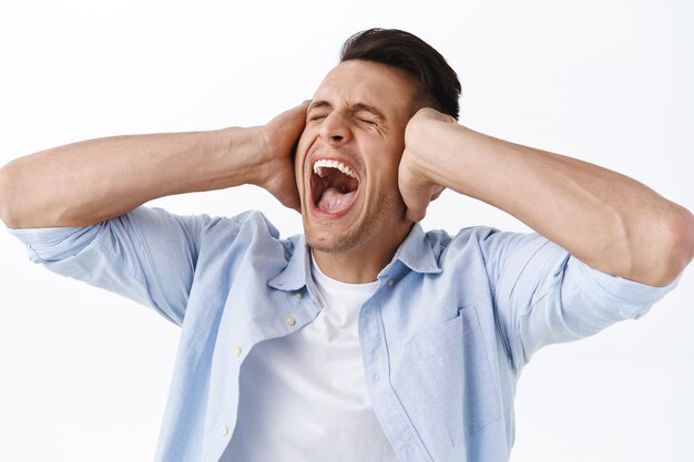 Close-up portrait of man screaming and shaking head in denial, close eyes and shut ears with hands, having emotional burnout on work, feel huge pressure and stressed, stand depressed white wall