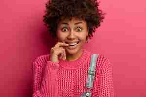 Free photo close up portrait of merry curious woman with curly afro hair, smiles gladfully , wears knitted oversized jumper, hears something hilarious and funny, isolated over rosy wall.