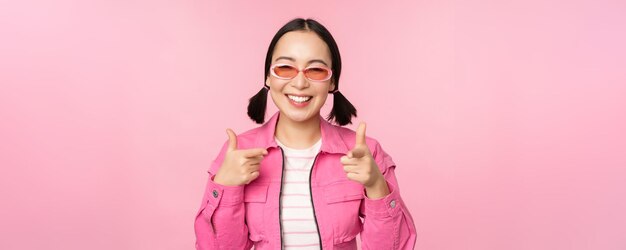 Крупный план портрета современной азиатской девушки в солнцезащитных очках, улыбающейся, указывающей пальцем на камеру, хвалит вас, приглашая или хваля, стоя на розовом фоне