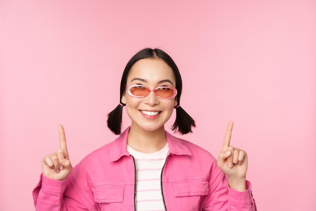 Close up portrait of modern korean female model wears sunglasses points fingers up shows advertisement promo banner pink background