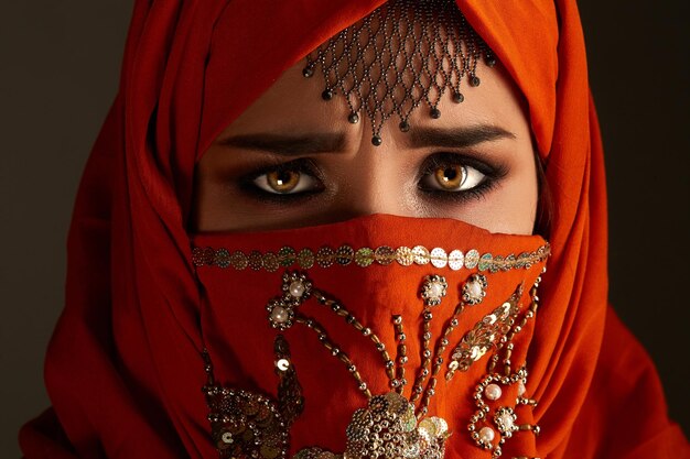 Close-up portrait of a moody young girl with charming smoky eyes and jewelry on the forehead, wearing the terracotta hijab decorated with sequins. She is posing and looking at the camera on a dark bac