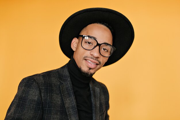 Close-up portrait of mulatto male model in black hat. Indoor photo of cheerful young man with brown skin.