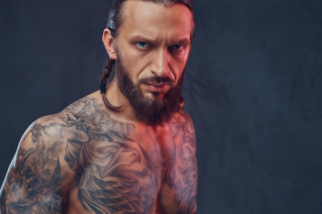 Close-up portrait of a muscular bearded tattoed male with a stylish haircut, isolated on a dark background.