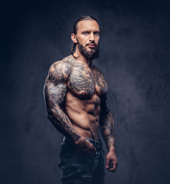 Крупный план мускулистого обнаженного бородатого мужчины с татуировками и стильной стрижкой. Изолированные на темном фоне.