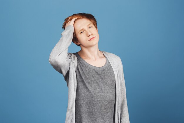 Close up portrait of narcissistic young good-looking guy with ginger hair in casual grey outfit fixing hair with hand,  with confident and flirty face expression.