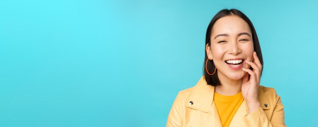 Крупный план портрета натуральной азиатской девушки, смеющейся, улыбающейся и выглядящей счастливой, стоящей на синем фоне