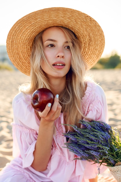 海の近くで週末を楽しんでいる麦わら帽子の自然な白人の女の子の肖像画を閉じます。果物でポーズをとる。ストローバッグのラベンダーの花束。