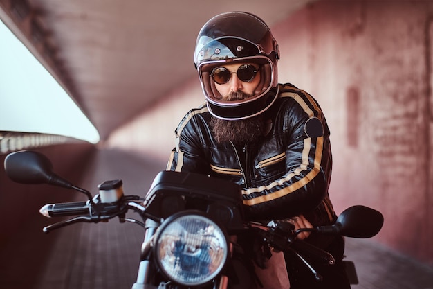 Бесплатное фото Крупный план брутального бородатого байкера в шлеме и солнцезащитных очках, одетого в черную кожаную куртку, сидящего на ретро-мотоцикле с включенной фарой.