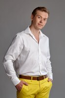 Крупным планом портрет молодого красивого рыжего парня в стильной белой рубашке и желтых брюках, смотрящего в камеру и улыбающегося во время позирования на сером студийном фоне. выражение лица человека. искренний