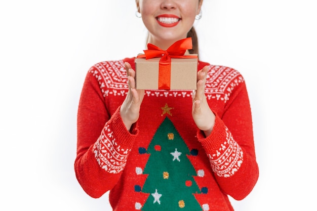 彼女の手に贈り物を持っている新年のセーターで若い笑顔の女性の肖像画を閉じます。白色の背景。クリスマスショッピングのコンセプト。