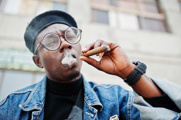 Бесплатное фото Крупный план портрета африканского американца в джинсовой куртке, берете и очках, курящего сигару