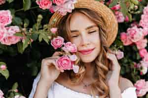 Бесплатное фото Портрет крупным планом экстатической фигурной девушки, позирующей с розами. открытый выстрел привлекательной женщины в соломенной шляпе, наслаждаясь летним днем.