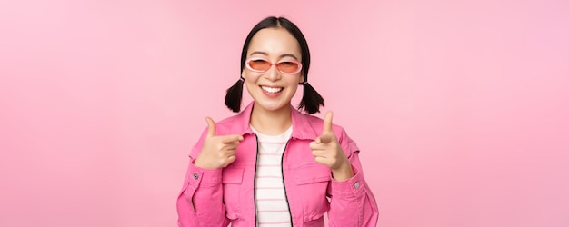 無料写真 サングラスをかけた現代アジアの女の子の肖像画をクローズアップカメラに指を指して笑ってピンクの背景の上に立ってあなたを招待または褒め称えます